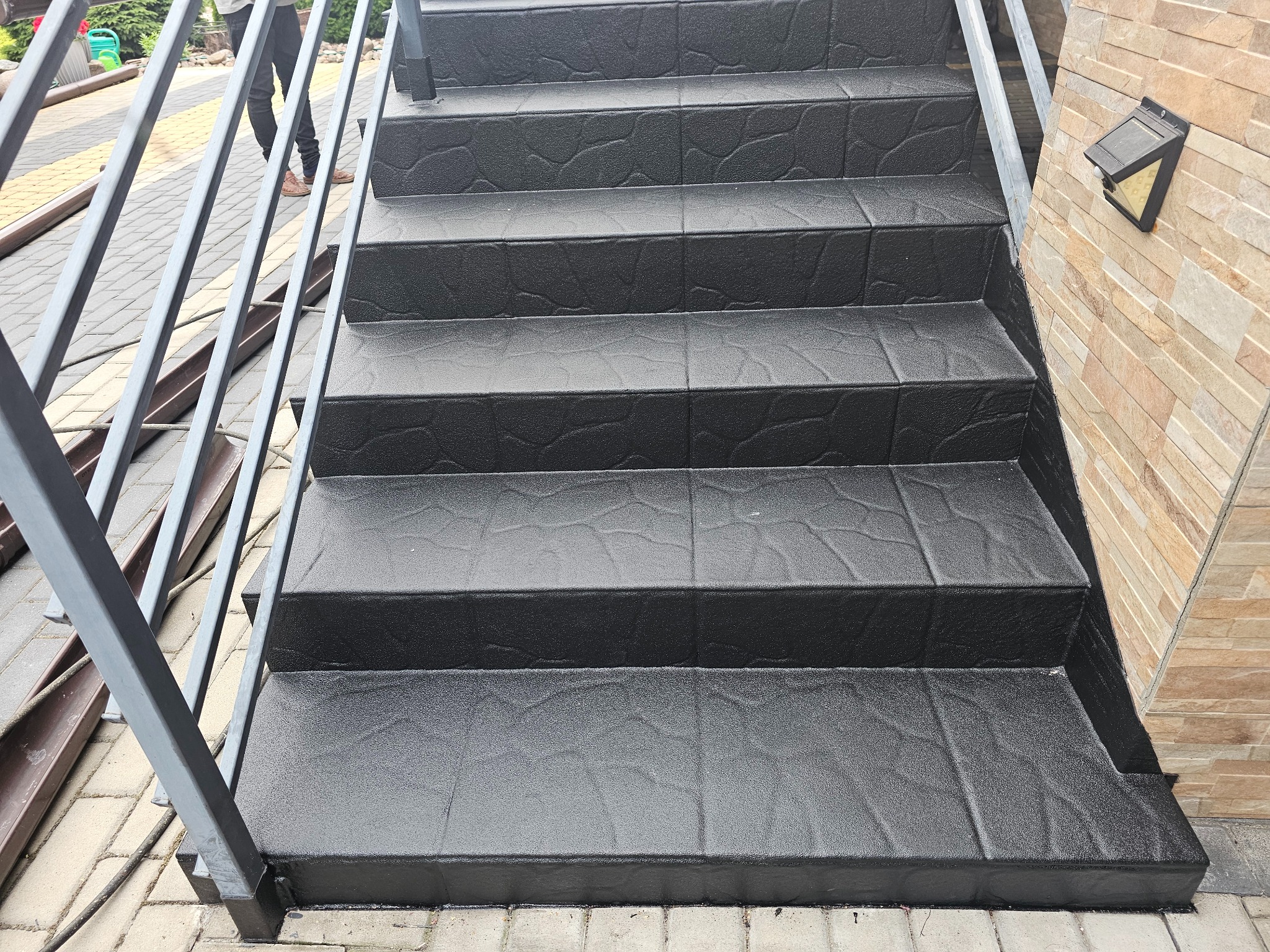 Schody z szarą, antypoślizgową posadzką żywiczną o fakturze kamienia, z metalową barierką. Widoczne oświetlenie zewnętrzne i fragment elewacji z cegły.