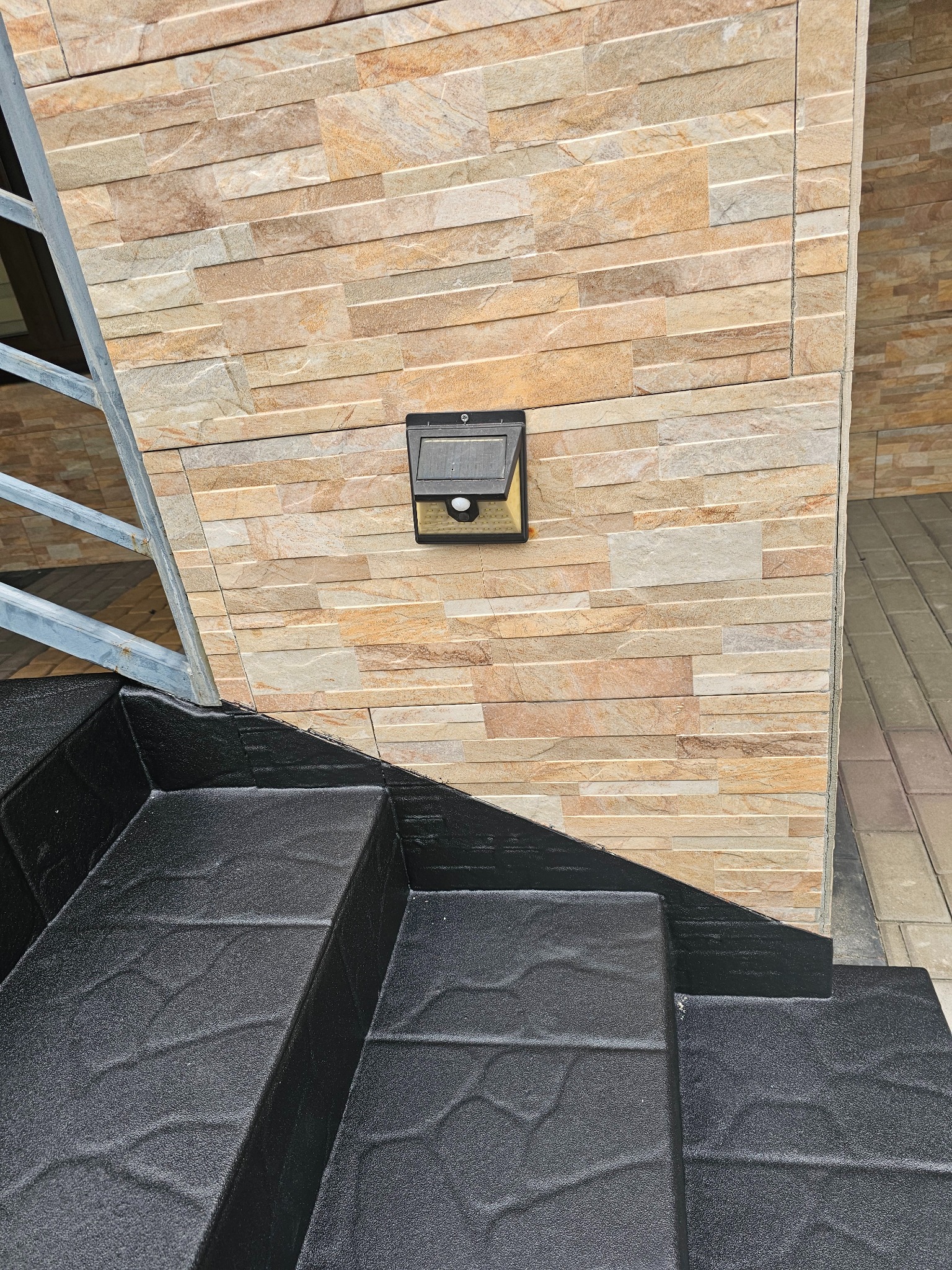 Czarne schody zewnętrzne z fakturą kamienia, pokryte jednolitą powłoką, kontrastują z elewacją z piaskowca i lampą solarną. Metalowa balustrada z lewej strony.