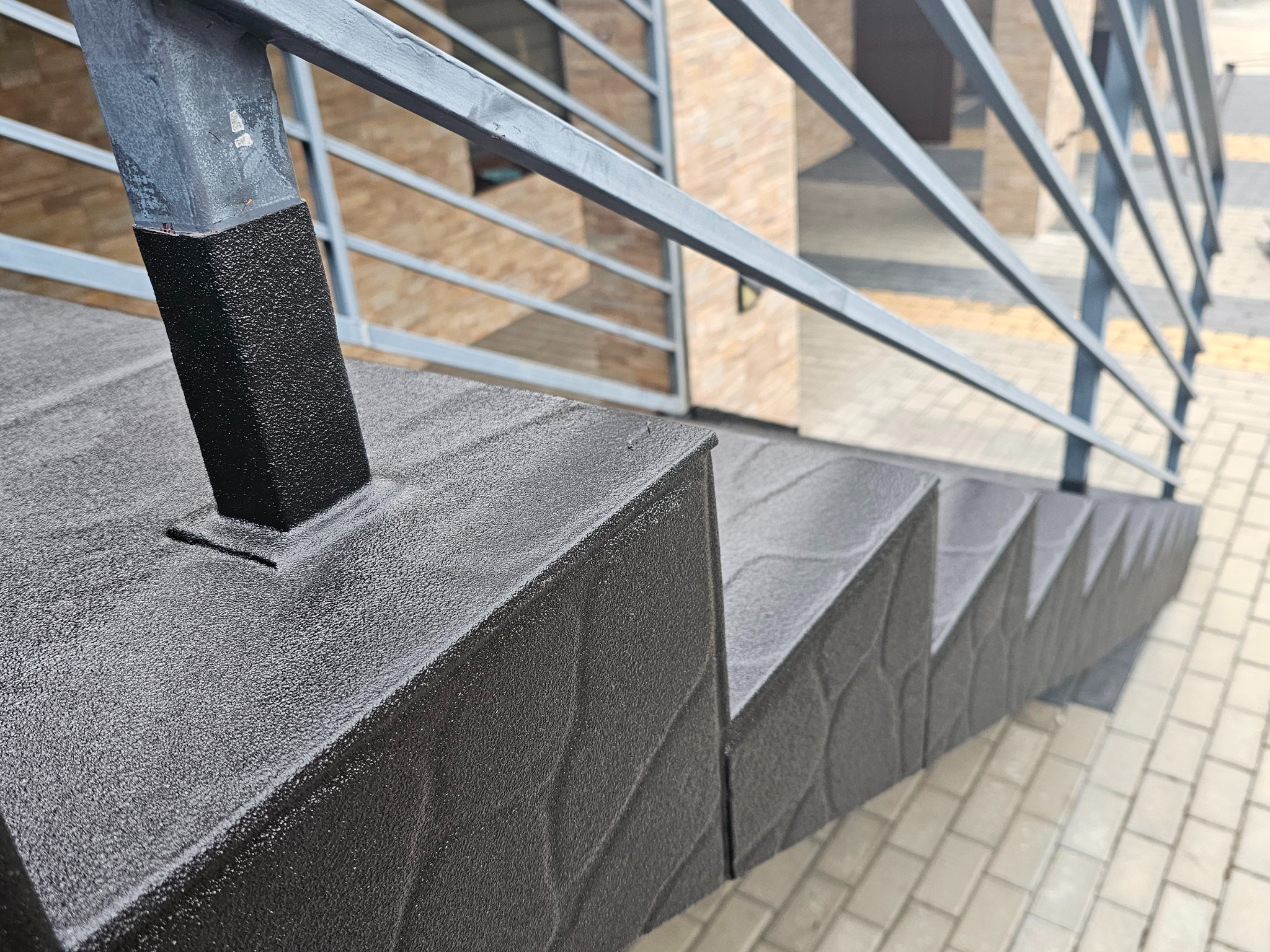 Schody zewnętrzne z szarą, chropowatą posadzką żywiczną i metalową barierką, widok z góry, faktura posadzki. Detal wykończenia.