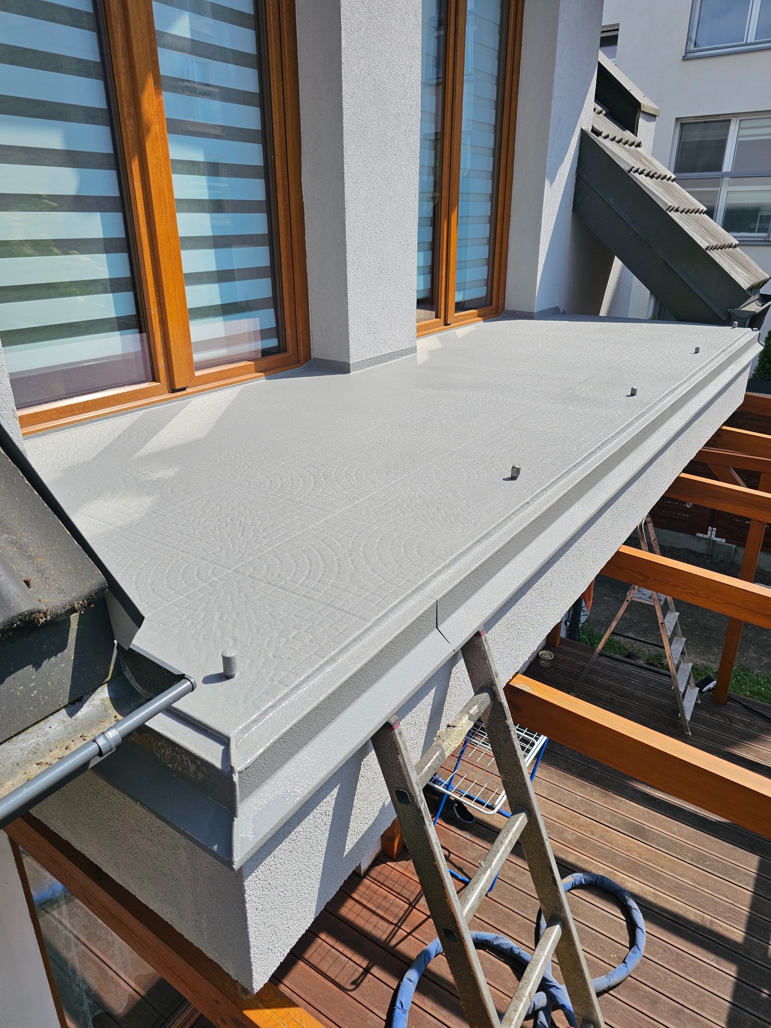 Szara, wzorzysta posadzka żywiczna na balkonie, widok z góry, z fragmentem drewnianej konstrukcji, drabiny i elewacji budynku. Detal wykończenia.