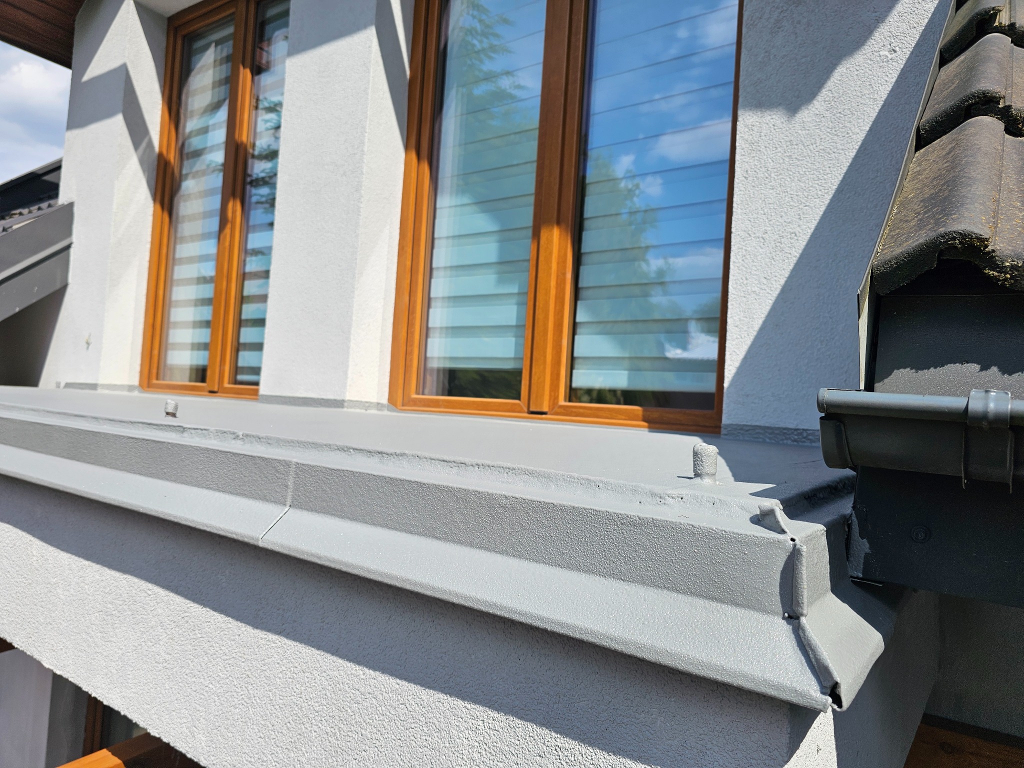 Szara, chropowata elewacja domu z drewnianymi oknami i widocznym fragmentem dachu z rynną. Detal wykończenia.