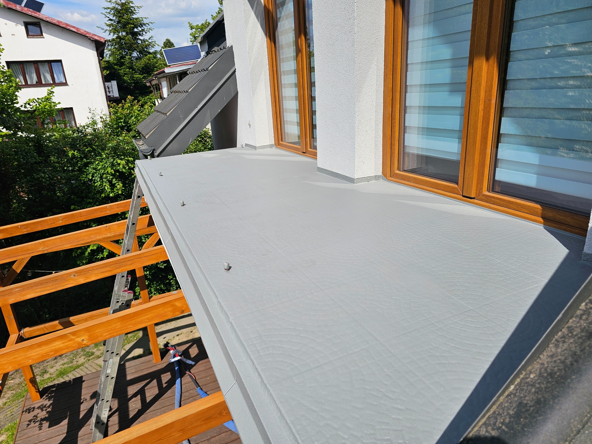 Szary, gładki dach z żywicy epoksydowej, detale okien z drewnianymi ramami i widoczna drabina. Nowoczesne wykończenie dachu z bliska, w słoneczny dzień.