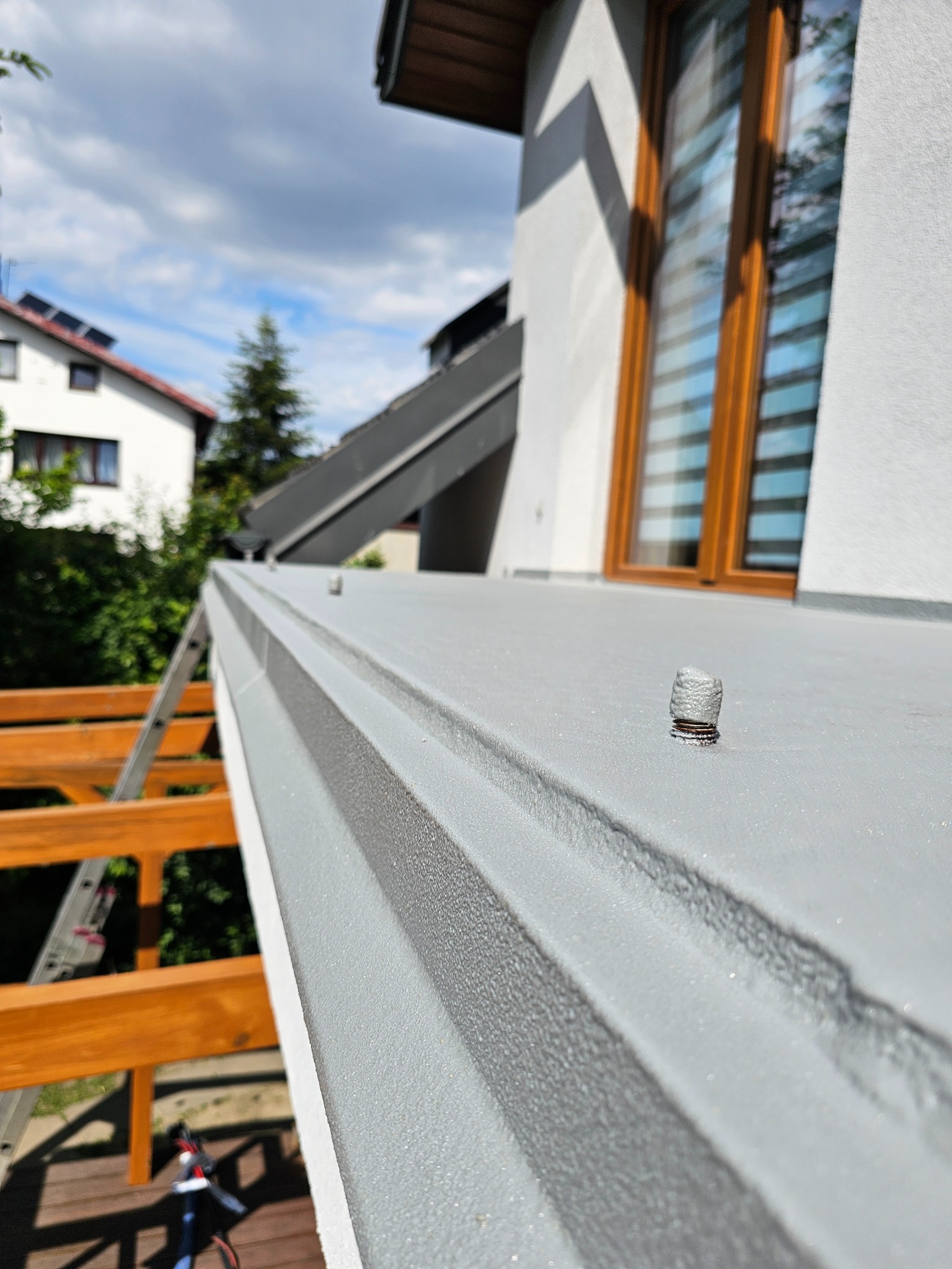 Szara, teksturowana powierzchnia z elementami mocującymi, prawdopodobnie pokryta natryskową izolacją. W tle okno domu i drewniana konstrukcja.