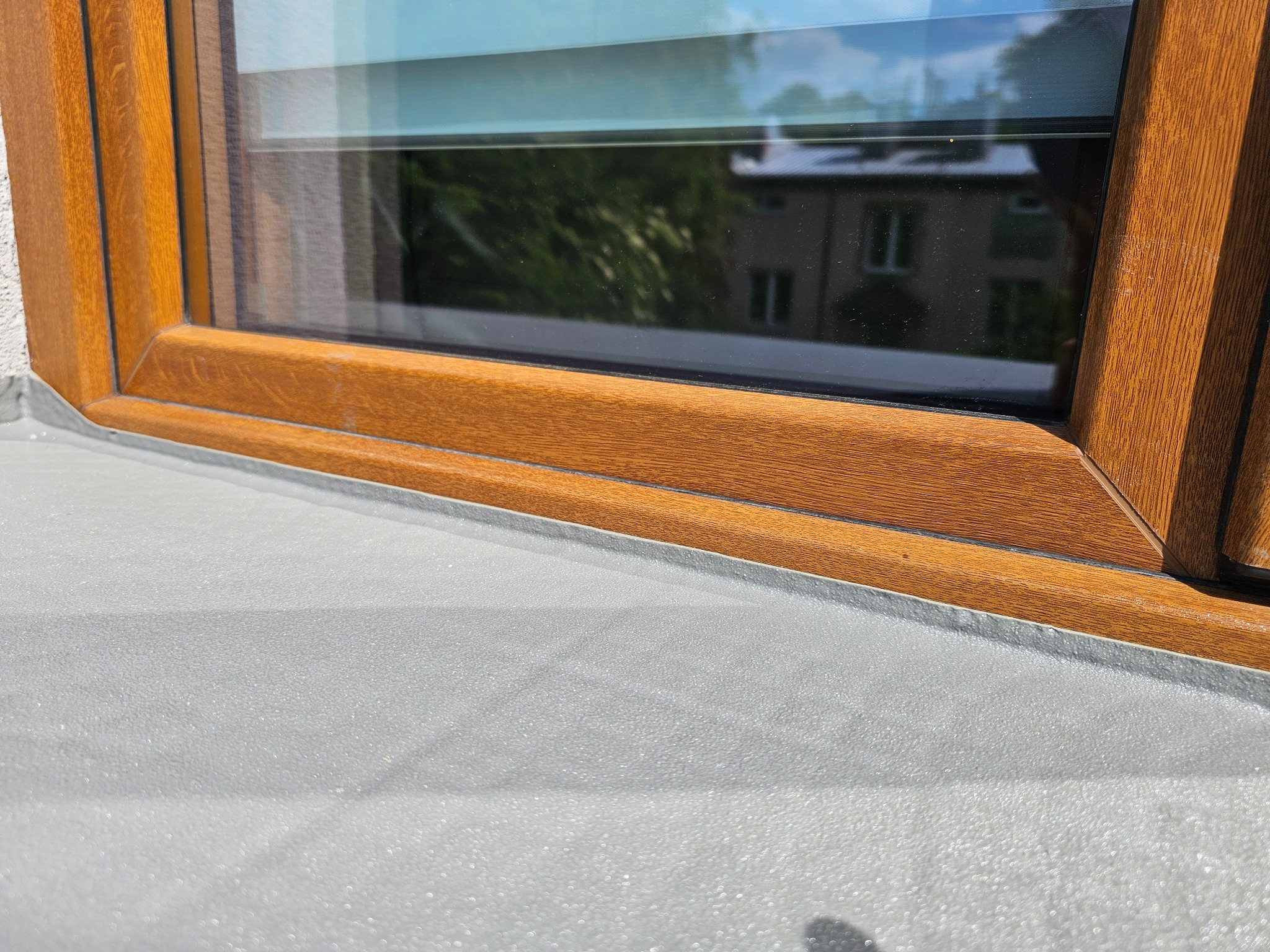 Szara posadzka żywiczna przy oknie z drewnianą ramą. Widoczne refleksy światła na gładkiej powierzchni posadzki. Okno odbija budynek i zieleń.