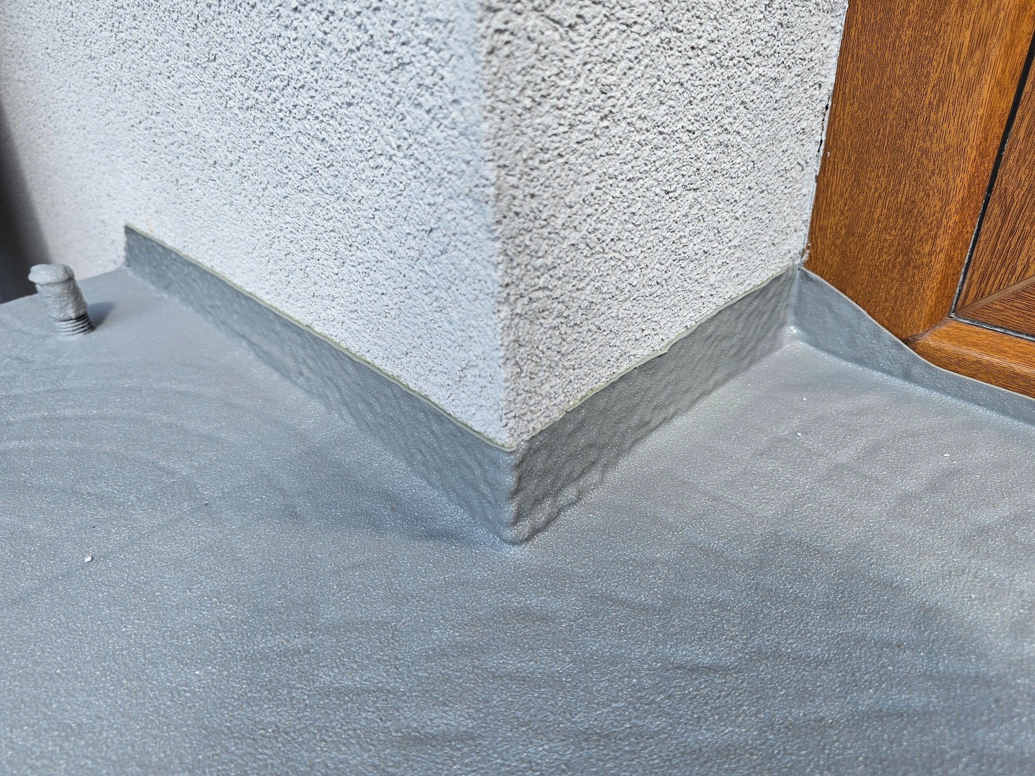 Szara, gładka posadzka żywiczna przy ścianie budynku z widocznym narożnikiem i fragmentem drzwi. Widoczne detale struktury posadzki i ściany.