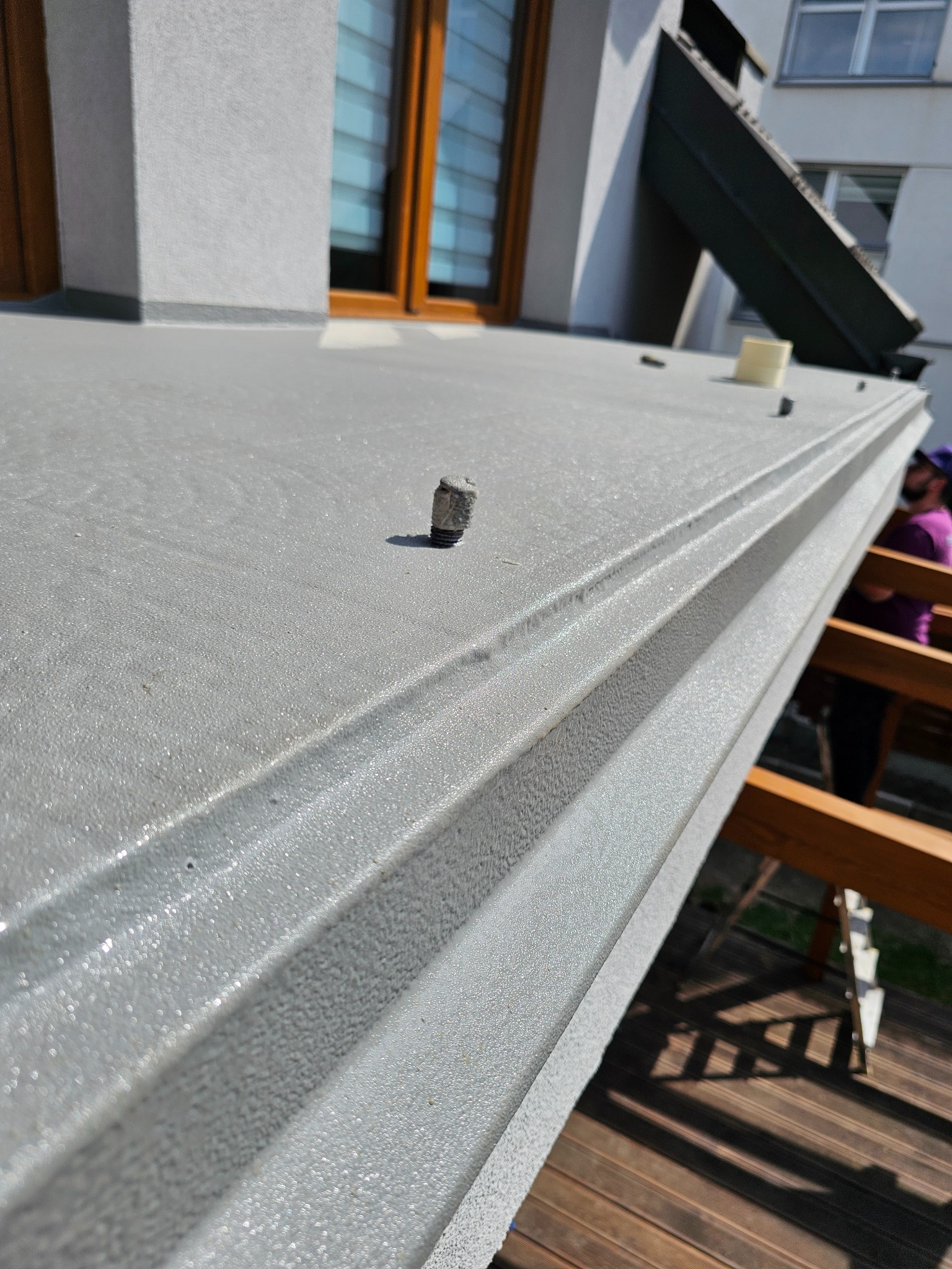 Szara, teksturowana powierzchnia z bliska, prawdopodobnie ocieplenie pianką PUR na dachu, z widocznymi elementami montażowymi i fragmentem rusztowania w tle.