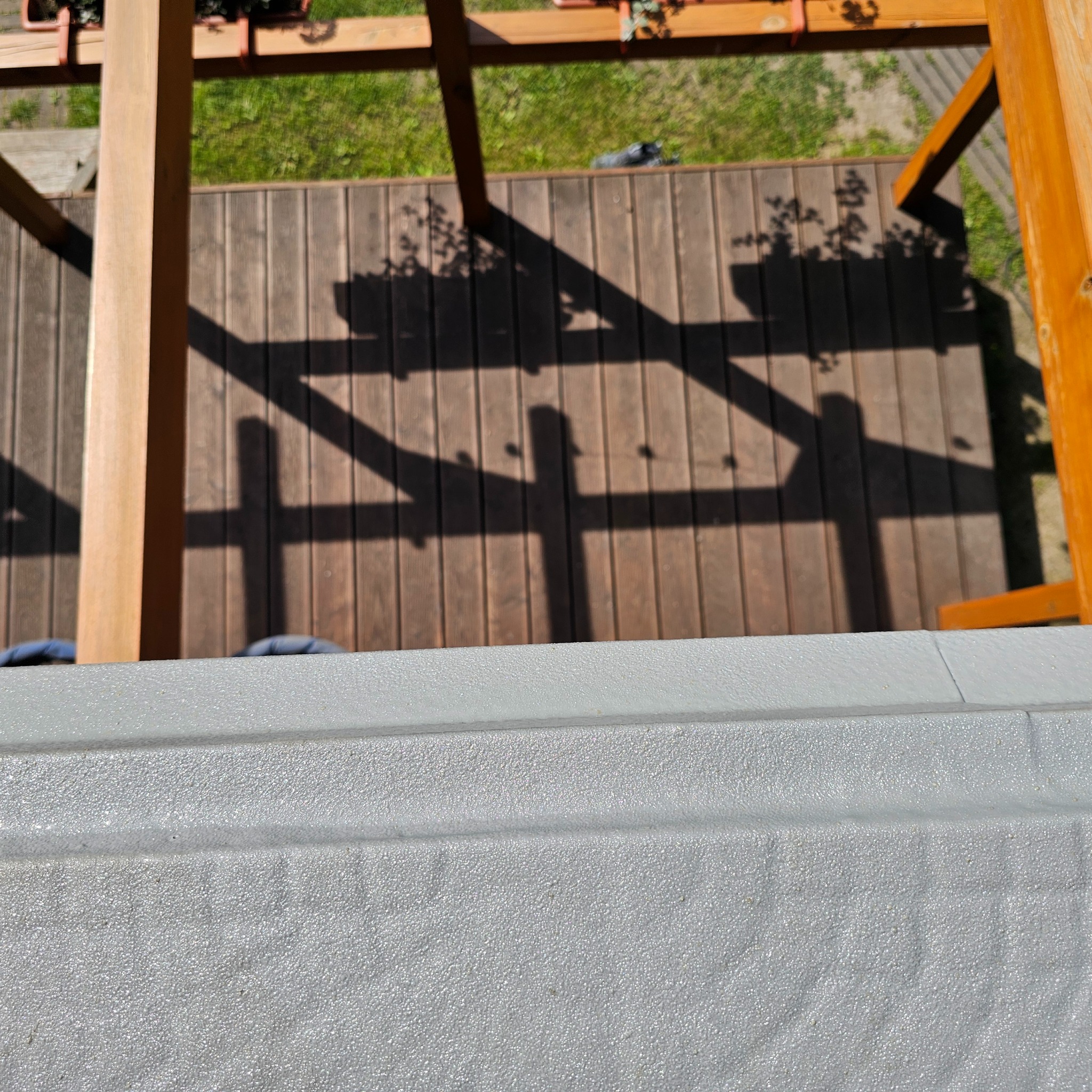 Szara, chropowata posadzka żywiczna na balkonie, z widokiem na drewniany taras i pergolę w słoneczny dzień. Detal faktury i cieni.