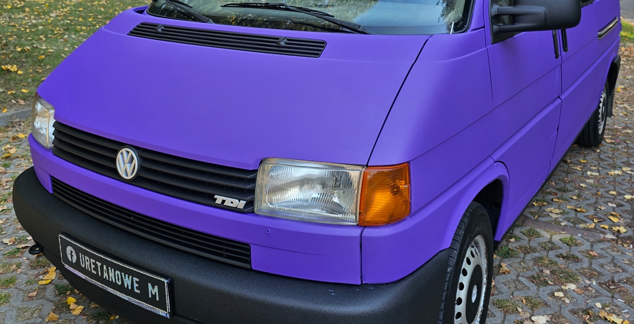 Fioletowy Volkswagen Transporter T4 po renowacji lakieru, z widocznym logo VW i tablicą rejestracyjną. Matowe wykończenie karoserii, jesienne tło.