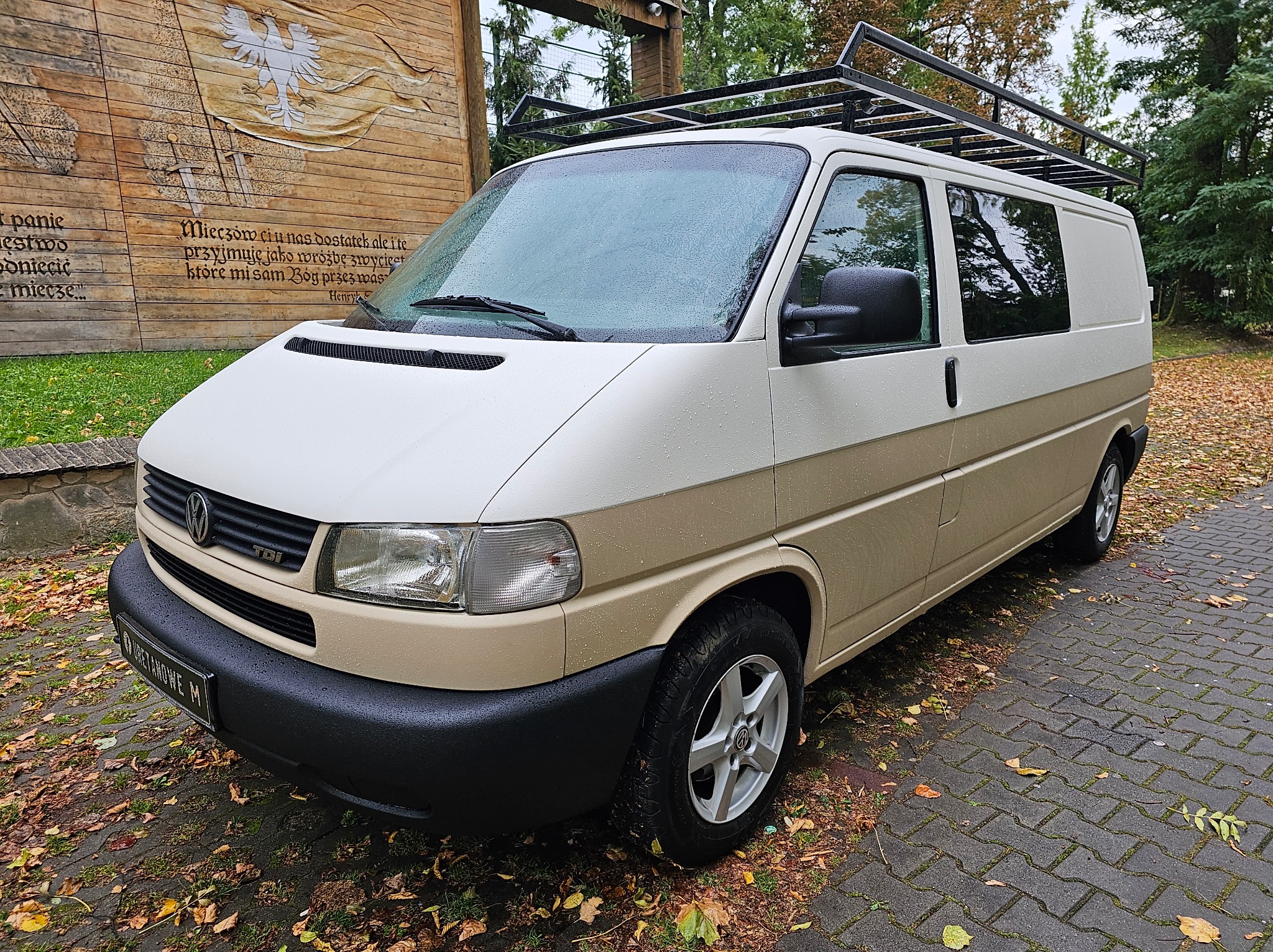 Dwukolorowy van VW Transporter T4 z bagażnikiem dachowym, zaparkowany na brukowanej nawierzchni z liśćmi, na tle drewnianej ściany z malowidłami patriotycznymi.
