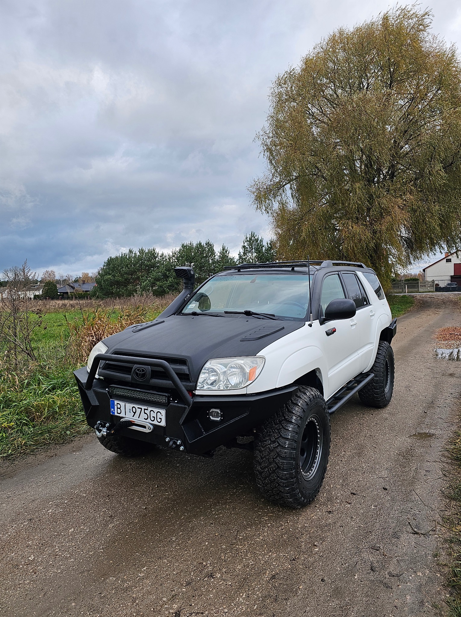 Biały terenowy SUV z czarnymi elementami (maska, orurowanie) na polnej drodze, z dużymi oponami i wyciągarką. W tle drzewo i zabudowania.