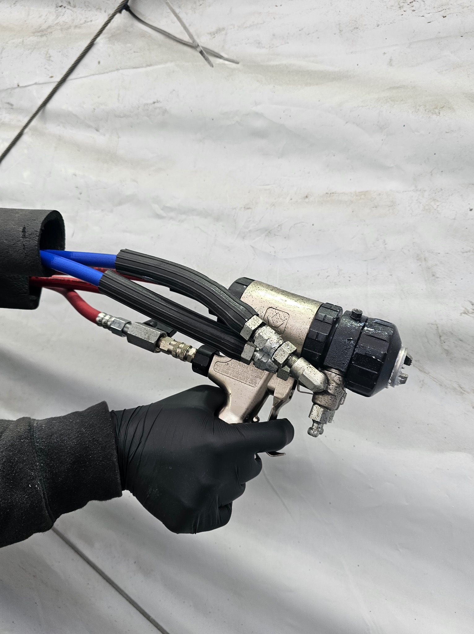 Pistolet natryskowy z przewodami (czerwony, niebieski) w dłoni w czarnej rękawicy, gotowy do aplikacji na białą powierzchnię. Detal urządzenia i węży.