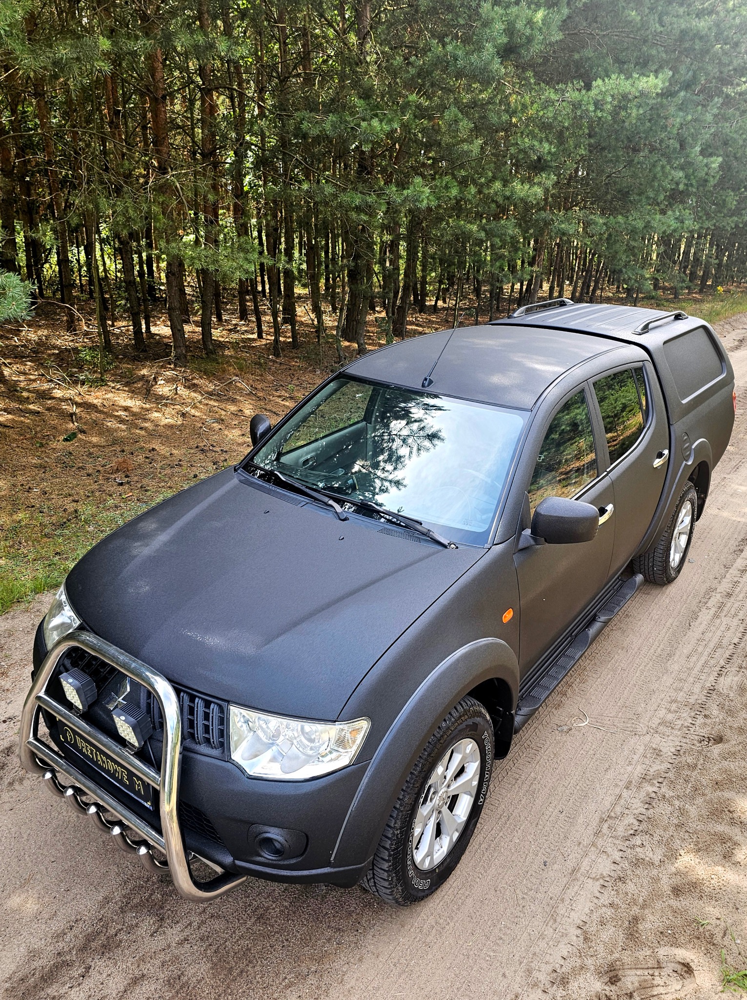 Czarny pickup z orurowaniem na tle leśnej drogi. Matowa powłoka karoserii, terenowe opony. Widoczne ślady opon na piasku i wysokie drzewa w tle.