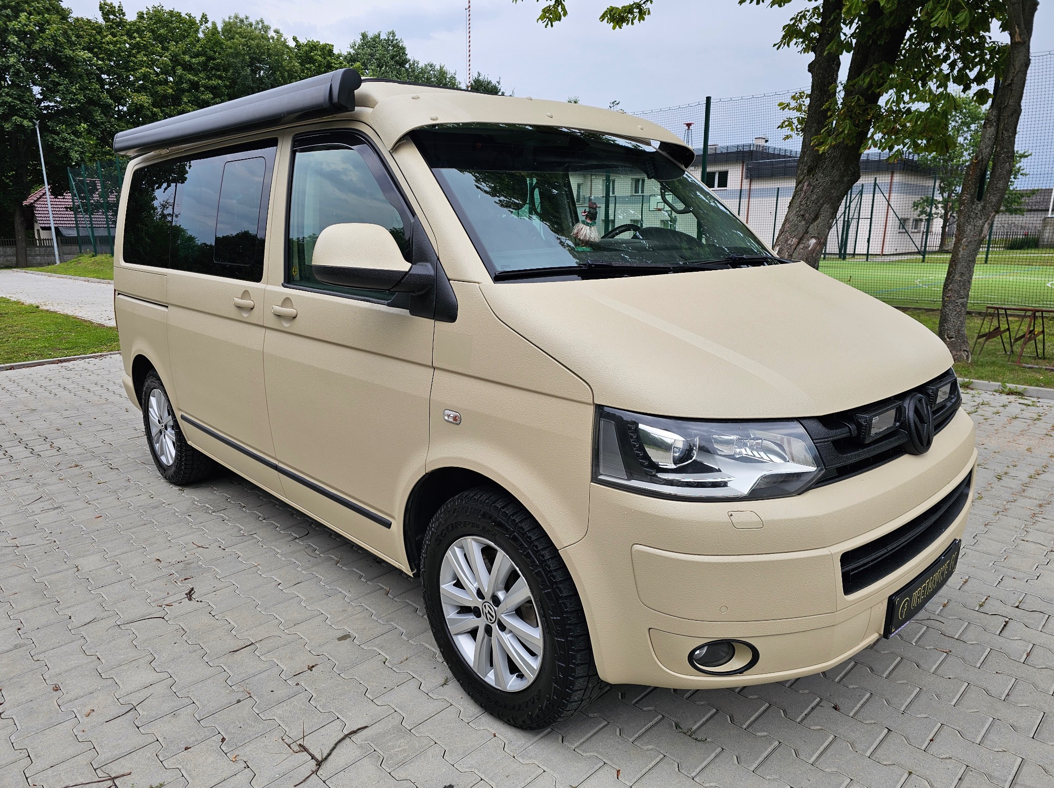 Beżowy van z teksturowaną powłoką ochronną, zaparkowany na brukowanej powierzchni. Widoczny markiza i osłona przeciwsłoneczna. Tło z zielenią i budynkiem.