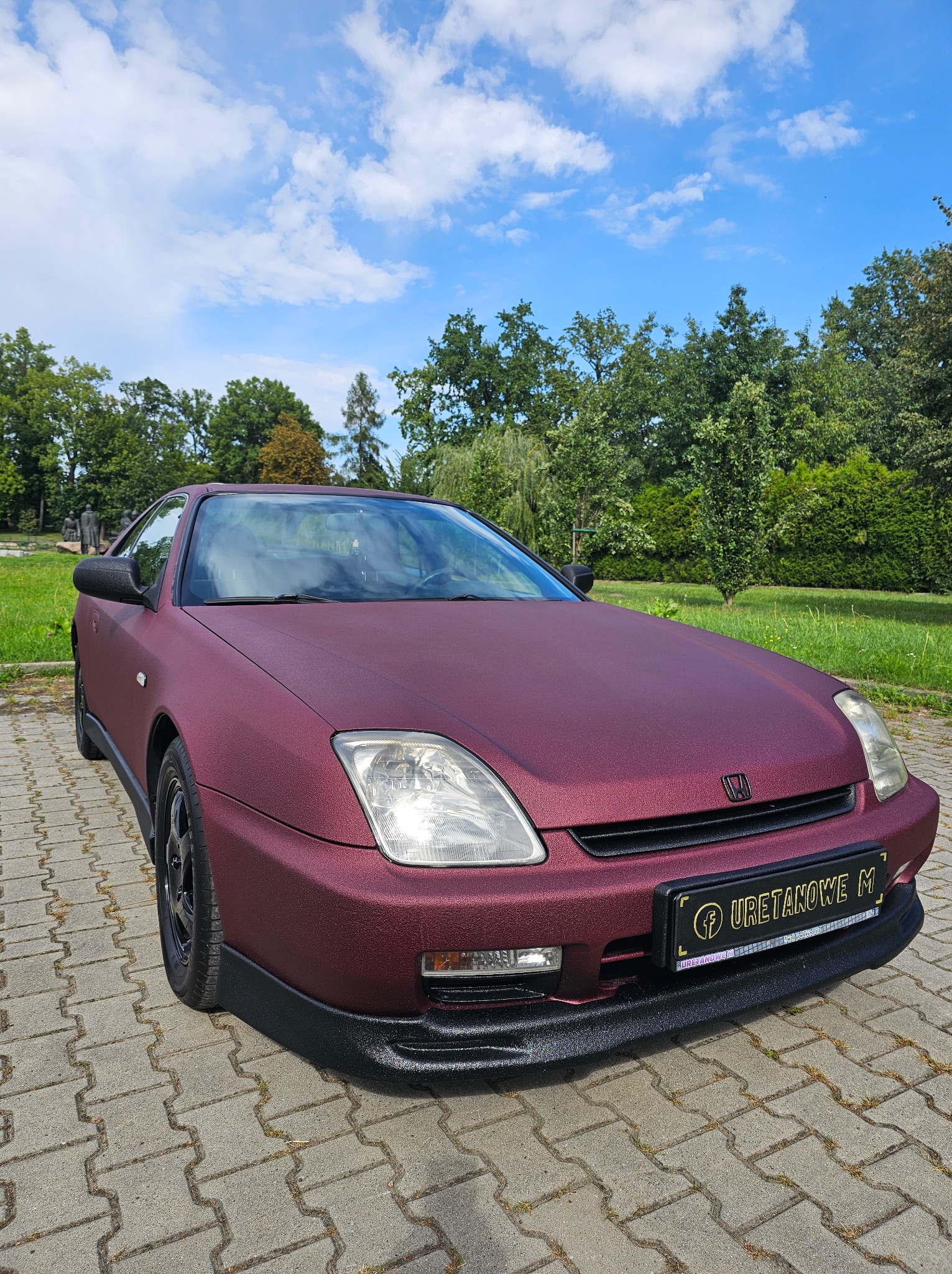 Bordowy samochód Honda Prelude z unikalną, teksturowaną powłoką, zaparkowany na kostce brukowej w otoczeniu zieleni i błękitnego nieba. Widoczna tablica z napisem 'URETANOWE M'.