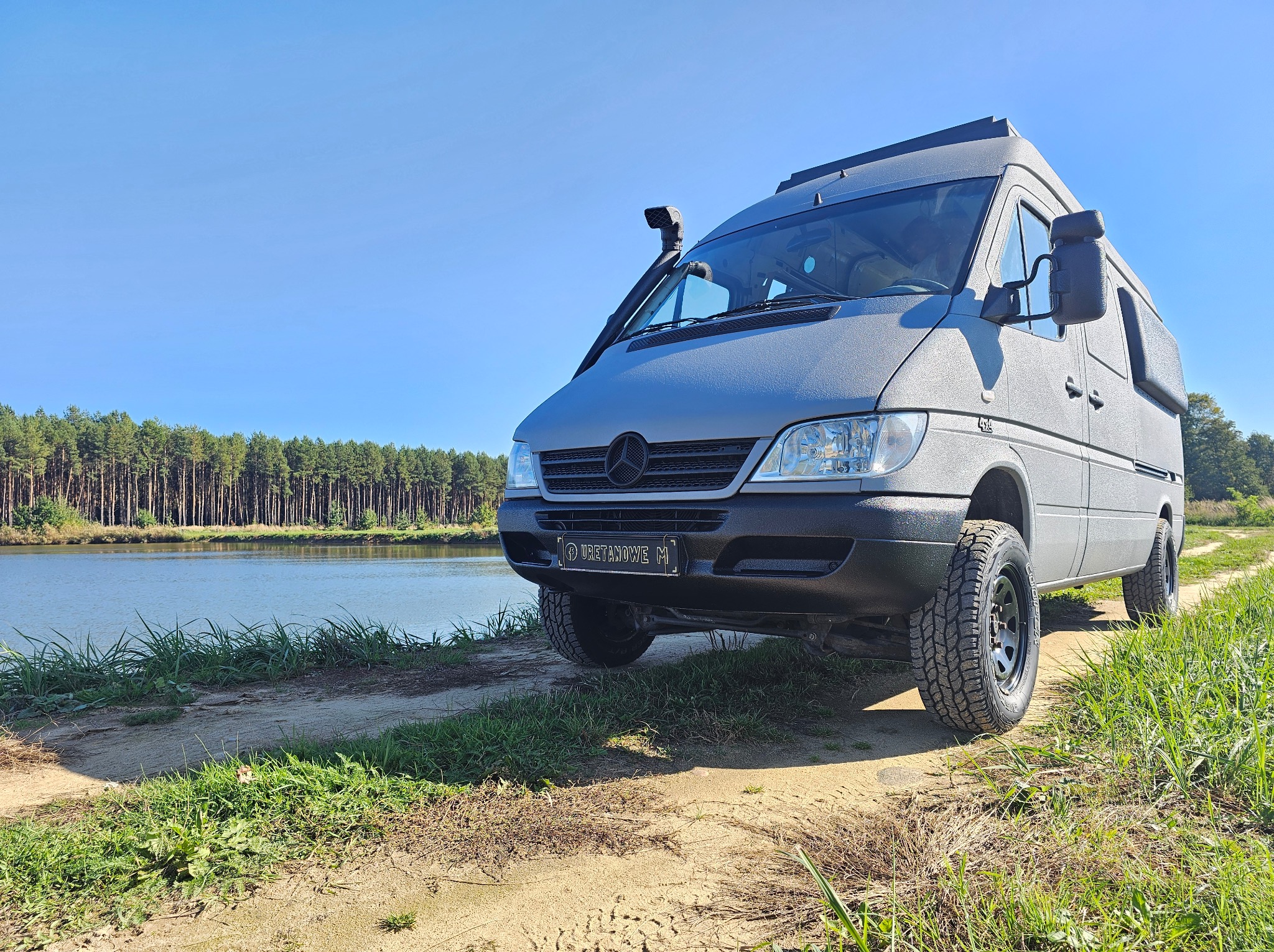 Szary van Mercedes Sprinter z chropowatą powłoką ochronną, snorkel i opony terenowe, zaparkowany na szutrowej drodze z widokiem na jezioro i las.