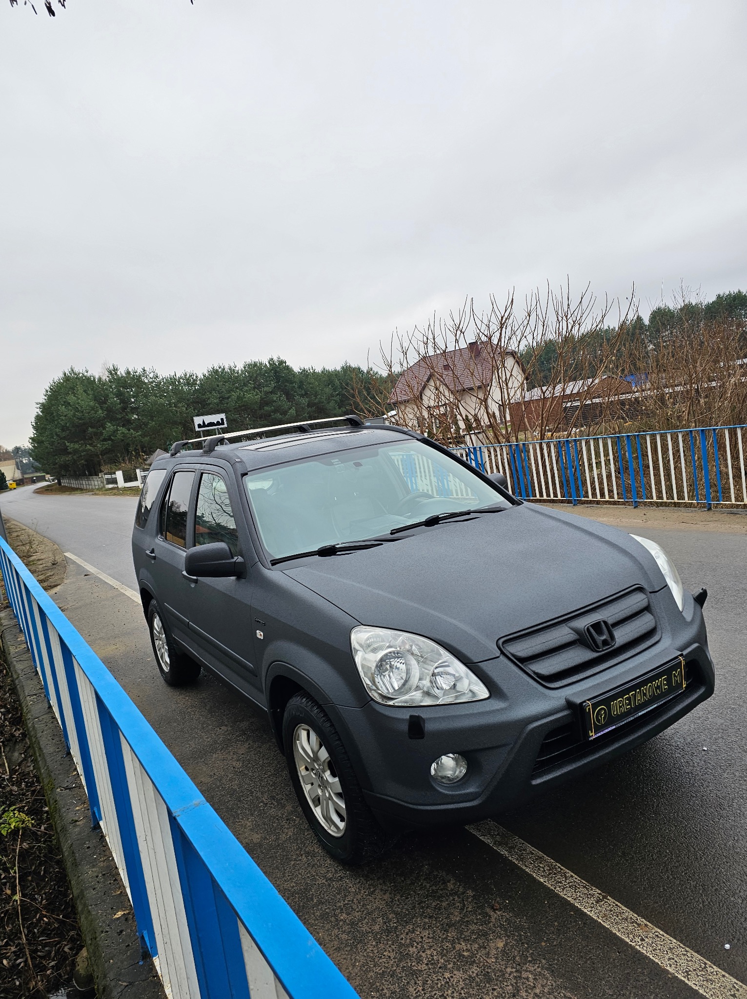 Szary SUV Honda CR-V z matowym wykończeniem, zaparkowany na mokrej drodze z niebiesko-białą barierką. W tle drzewa i dom. Tablica rejestracyjna z napisem 'Uretanowe M'.