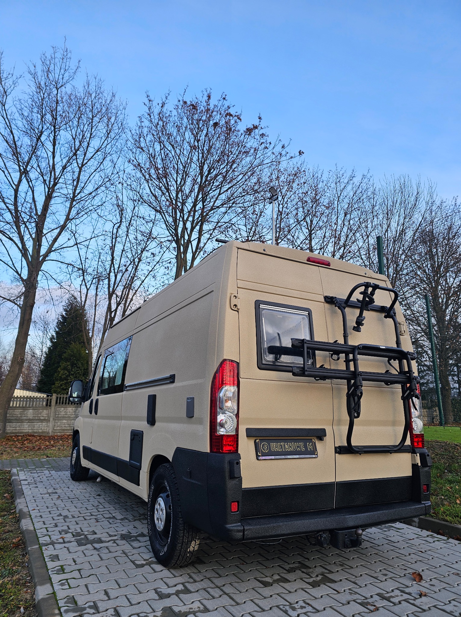 Beżowy kamper z bagażnikiem rowerowym, pokryty natryskową powłoką poliuretanową, widoczny na zewnątrz w pochmurny dzień. Teksturowana powierzchnia.