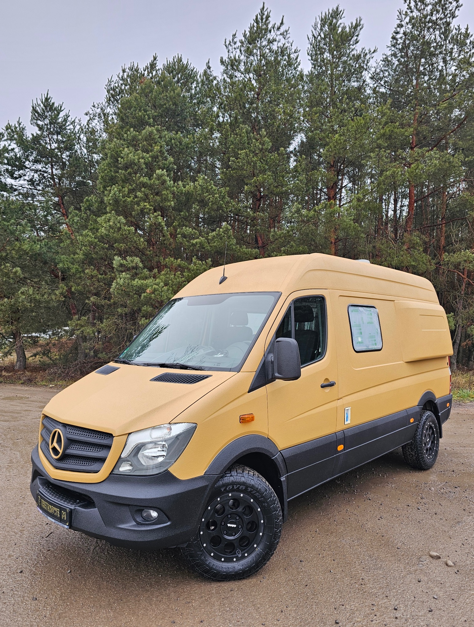 Żółty kamper Mercedes Sprinter z powłoką ochronną, terenowe opony, na tle lasu. Wykończenie odporne na zarysowania i warunki atmosferyczne.