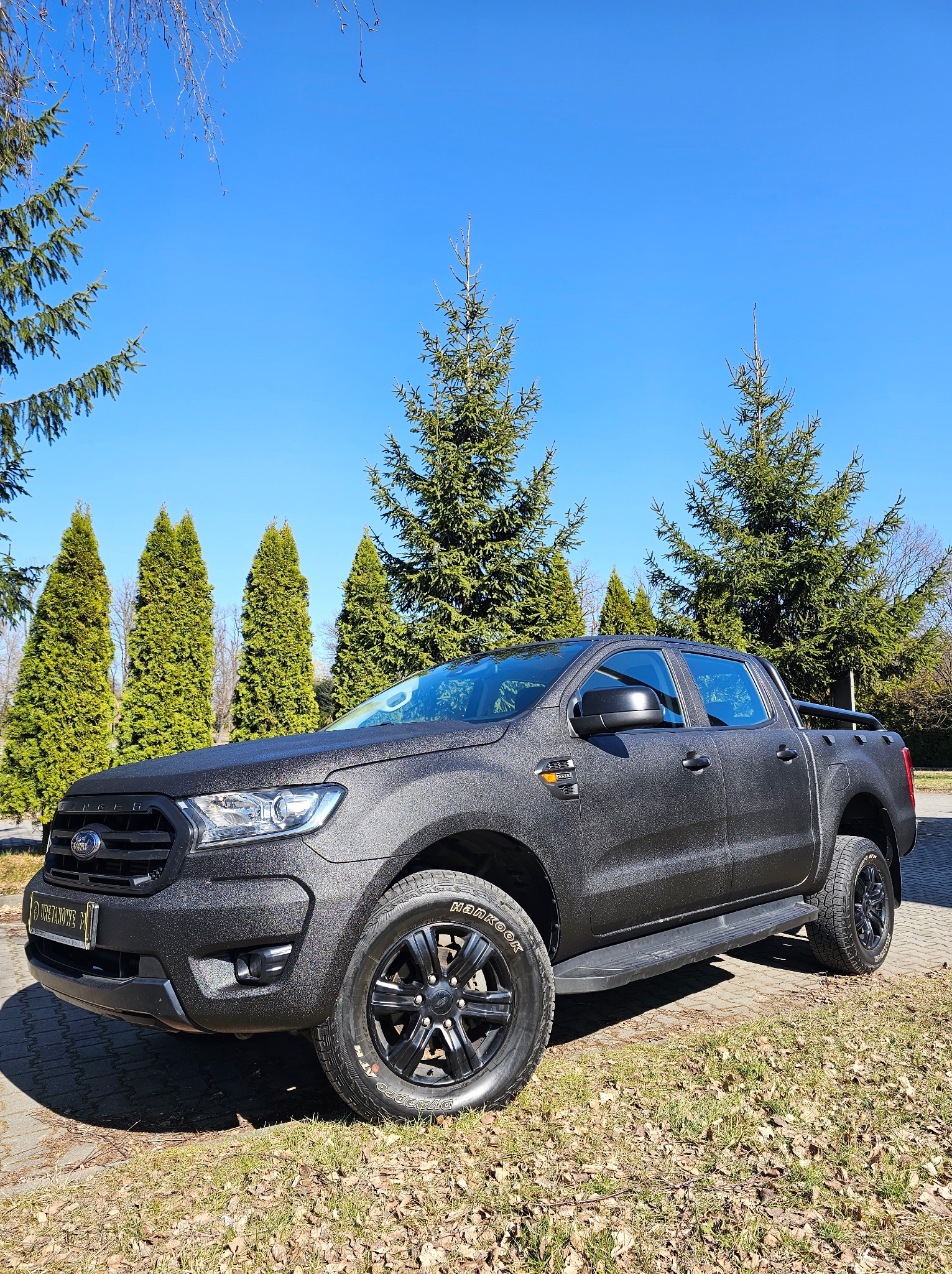 Czarny Ford Ranger po aplikacji ochronnej powłoki z fakturą, na tle zielonych drzew i błękitnego nieba. Widok z boku, podkreślający strukturę i trwałość zabezpieczenia.