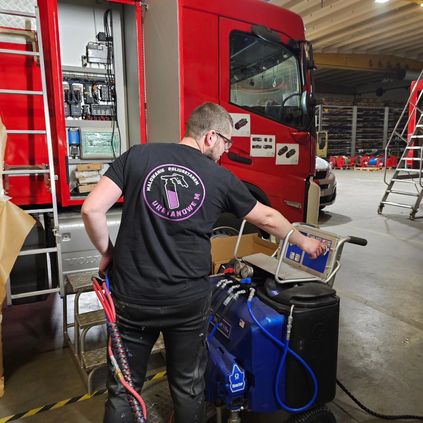 Pracownik w czarnej koszulce z logo, obsługuje niebieskie urządzenie natryskowe w hali, prawdopodobnie podczas aplikacji pianki poliuretanowej na element pojazdu.