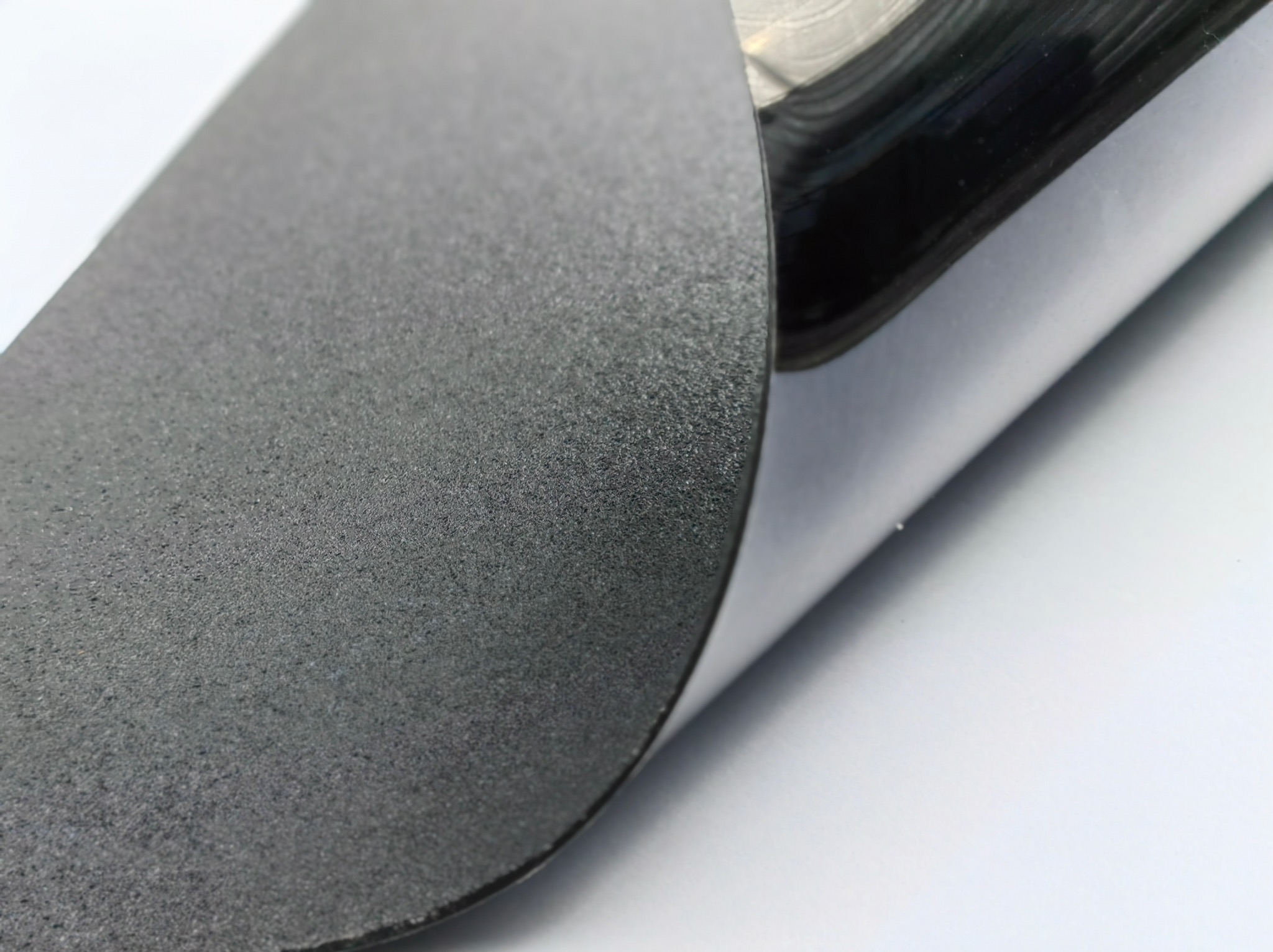 Zbliżenie na czarną, teksturowaną powierzchnię z podwiniętym rogiem, ukazującą gładką, srebrną warstwę spodnią. Detal faktury i kontrast materiałów.