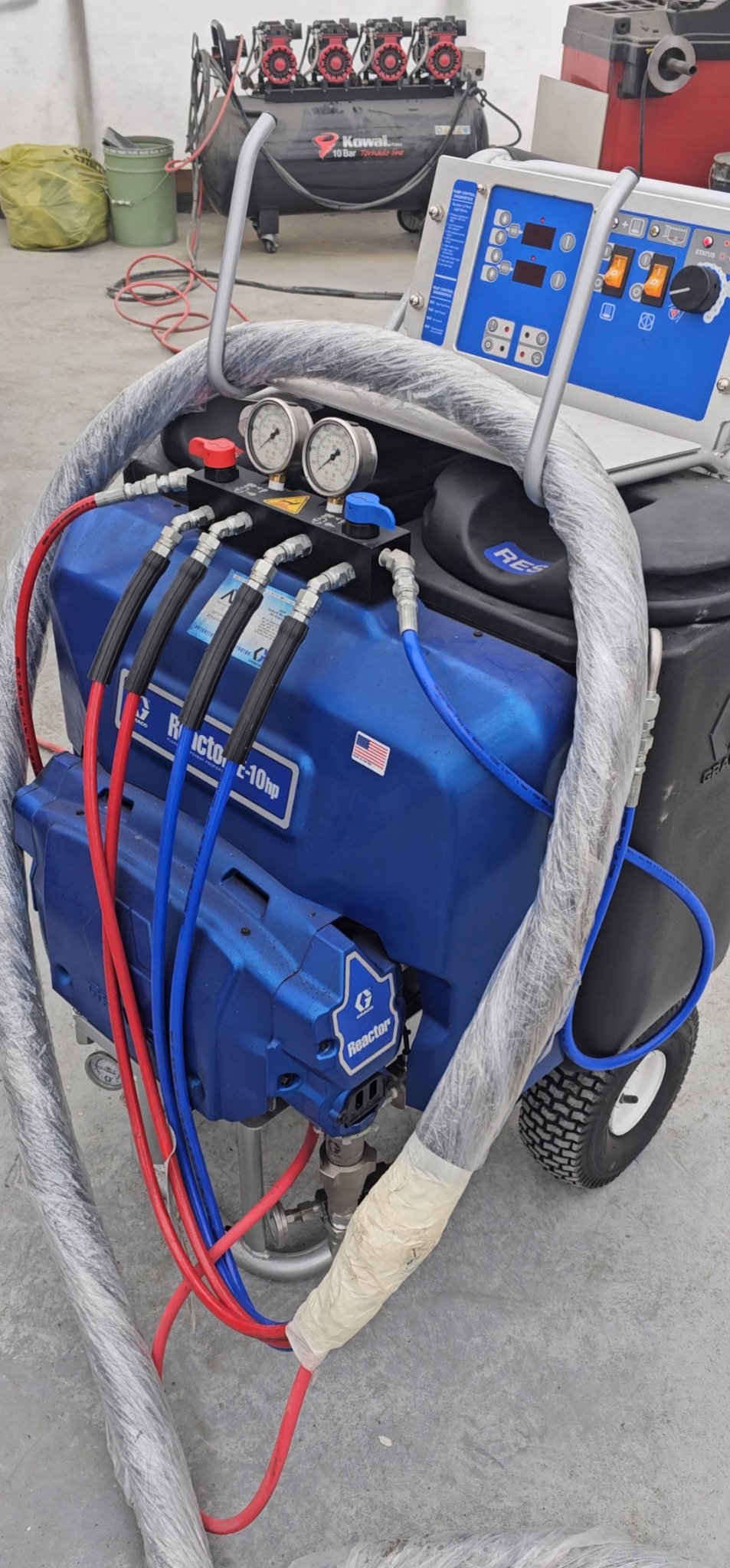 Niebieski agregat natryskowy Reactor E-10hp na wózku z manometrami i przewodami w osłonie. W tle kompresor Kowal. Sprzęt do natrysku pianki PUR lub posadzek żywicznych.