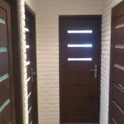 Korytarz z trzema ciemnymi drzwiami na tle białej ściany z cegły, wykończony panelami podłogowymi imitującymi drewno.