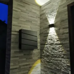 Ściana wyłożona szarą cegłą dekoracyjną, oświetlona górnym punktowym światłem i kinkietem rzucającym stożki światła w górę i w dół, obok czarnej, poziomo żebrowanej skrzynki.