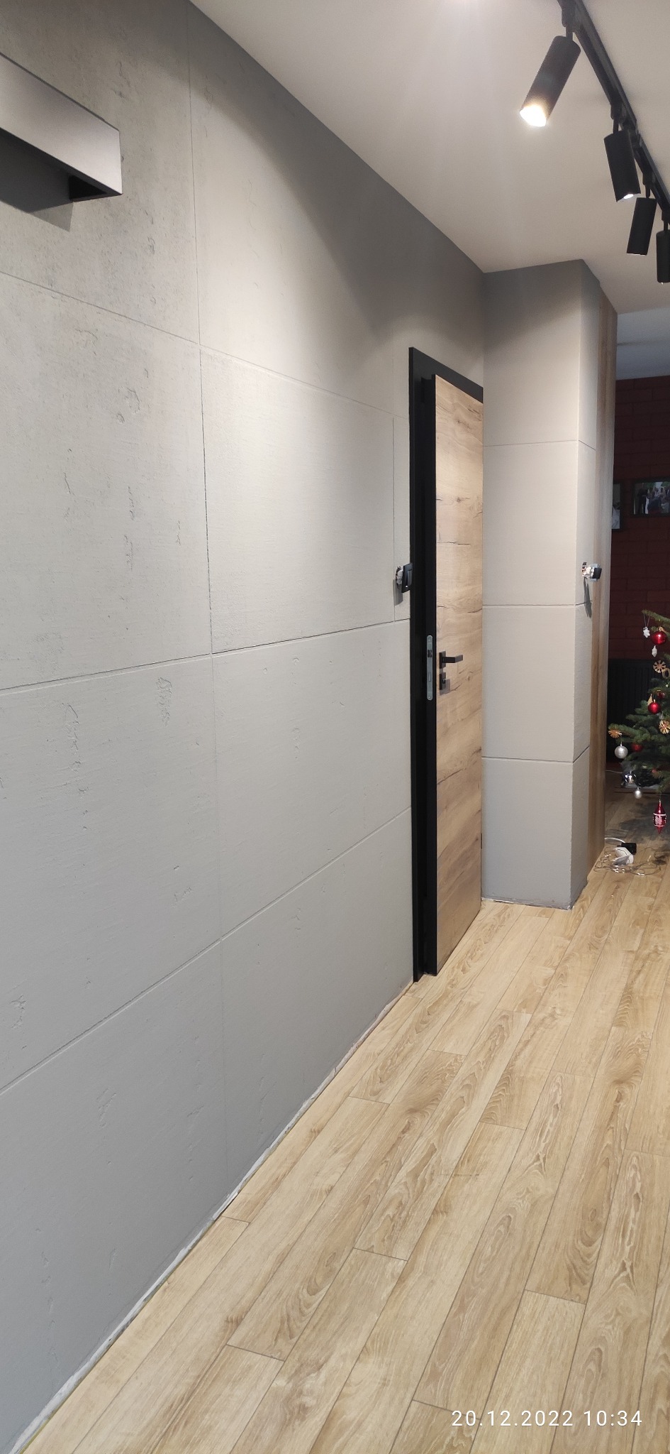 Wnętrze po remoncie: szara ściana z płyt imitujących beton, drewnopodobna podłoga, drzwi wewnętrzne z czarną ościeżnicą i drewnianym skrzydłem, oświetlenie punktowe na suficie, choinka w tle.
