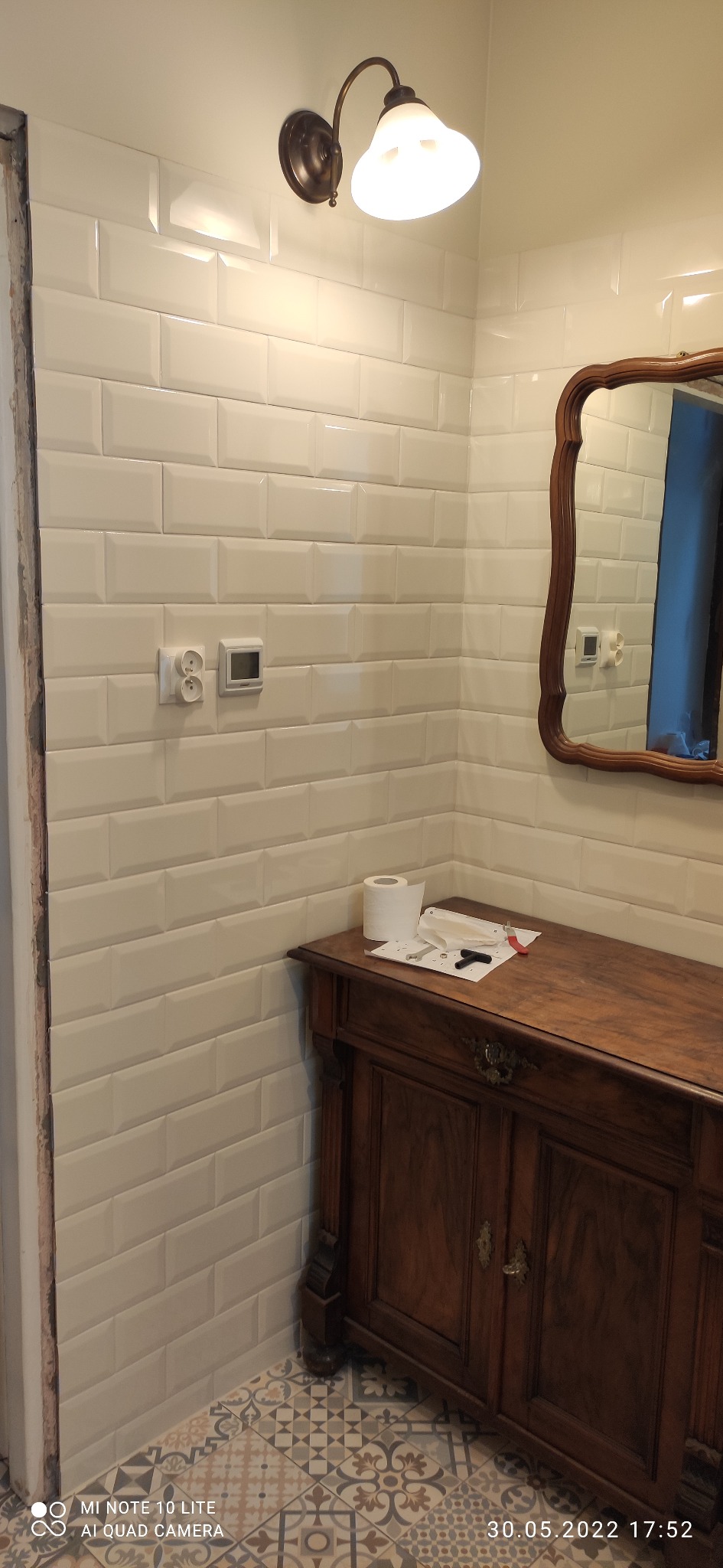 Ściana w łazience wyłożona białymi, prostokątnymi płytkami, z widocznymi gniazdkami elektrycznymi i termostatem, nad drewnianą komodą z akcesoriami łazienkowymi i lustrem w ozdobnej ramie.