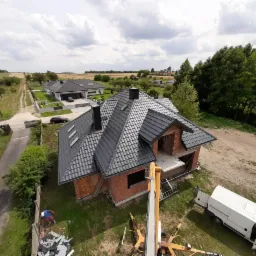 Montaż grafitowej dachówki na nowym domu w trakcie budowy, widok z podnośnika koszowego, w tle inne domy i pola uprawne.