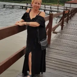 Kobieta w czarnej sukience na drewnianym pomoście w Dobrodzieniu. Sukienka z falbanami i rozcięciem, torba na ramię. W tle jezioro i las.