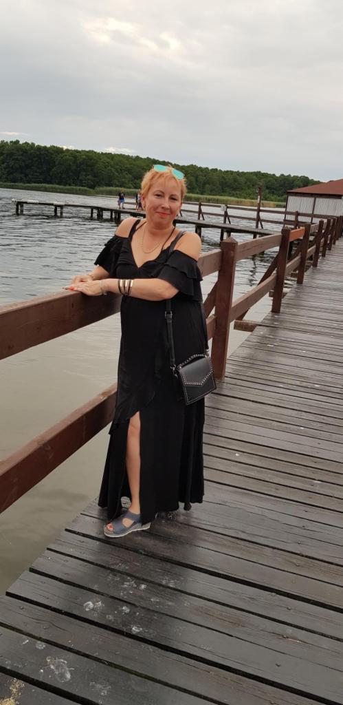 Kobieta w czarnej sukience na drewnianym pomoście w Dobrodzieniu. Sukienka z falbanami i rozcięciem, torba na ramię. W tle jezioro i las.