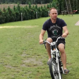 Mężczyzna na elektrycznym rowerze na trawniku, w tle tuje i plac zabaw. Nietypowy rower z grubymi oponami i widocznym silnikiem.