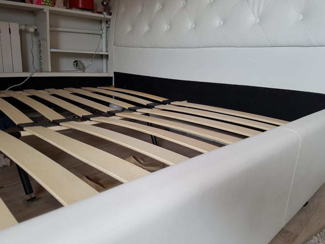 Rozłożone łóżko z białą ramą i drewnianym stelażem, widoczne elementy montażowe i fragment szafki w tle.