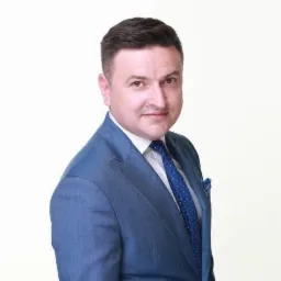 Portret uśmiechniętego mężczyzny w niebieskim garniturze i krawacie w kropki, z poszetką w kieszeni, na jasnym tle.