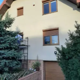 Montaż brązowych rolet zewnętrznych na elewacji domu jednorodzinnego, widok z dołu na rusztowanie i pracownika przy oknie.