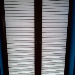 Zamknięte, poziome, białe żaluzje drewniane w ciemnobrązowej ramie okiennej, na tle błękitnej ściany.