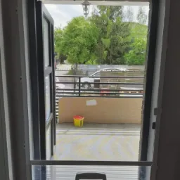 Moskitiera w otwartych drzwiach balkonowych z zamontowanym przejściem dla zwierząt, widok na balkon i zieleń na zewnątrz.