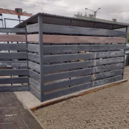 Nowoczesna osłona na pojemniki na śmieci z szarego metalu i elementami w kolorze drewna, częściowo owinięta folią ochronną, stojąca na betonowej kostce brukowej.