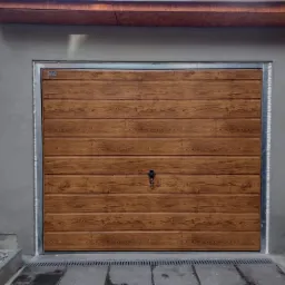 Zewnętrzna brama garażowa segmentowa z paneli imitujących drewno, z widocznym zamkiem, osadzona w surowej, szarej elewacji budynku z widocznym zadaszeniem i obrzeżem z metalu