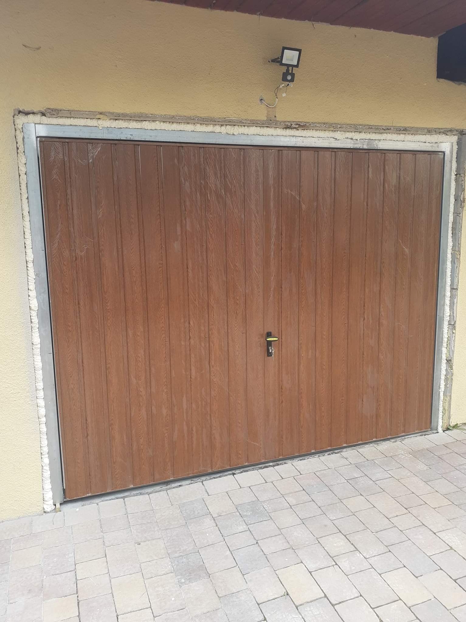 Brązowa brama garażowa z imitacją drewna, z metalową ramą, zamontowana w żółtej ścianie budynku, z widoczną kostką brukową na podjeździe.