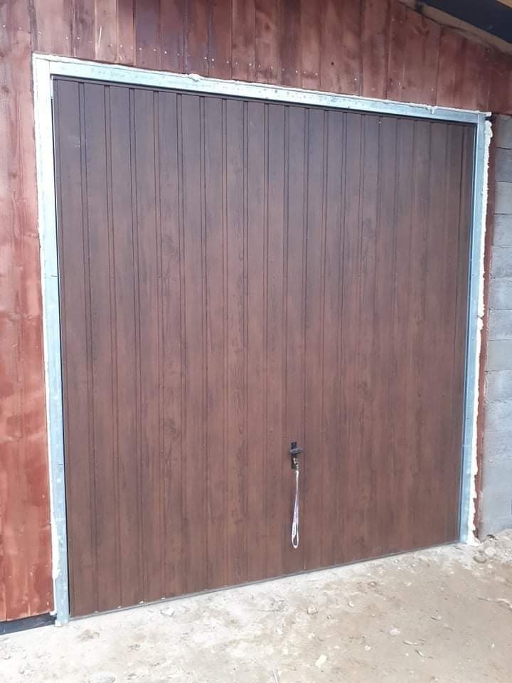 Zamknięte, brązowe, blaszane drzwi garażowe z metalową ramą, widoczne z zewnątrz, zamek z zawieszką, na tle drewnianej ściany i surowego betonu.