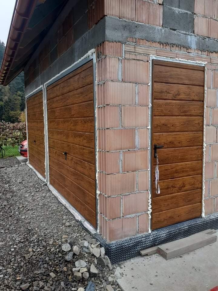 Niedokończona budowa garażu z dwoma bramami i drzwiami wejściowymi, z widocznymi cegłami i pianką montażową, teren wokół wysypany kamieniami.