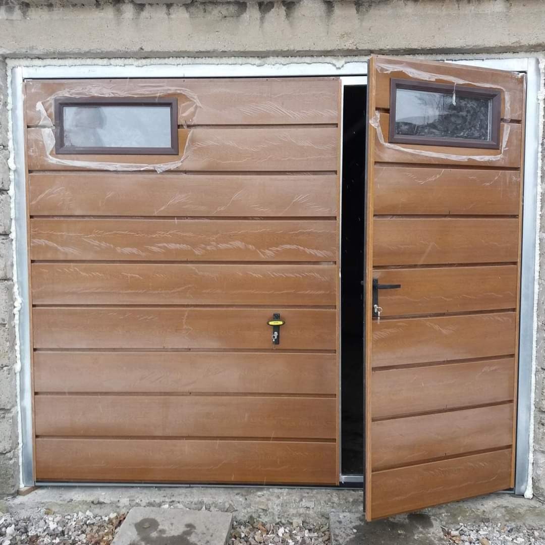 Brązowa, segmentowa brama garażowa z dwoma małymi oknami, częściowo otwarta i zabezpieczona folią ochronną.