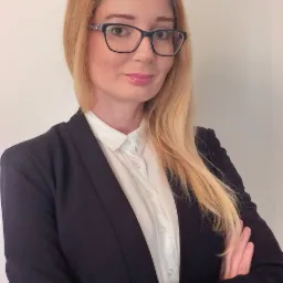 Portret kobiety w okularach, białej koszuli i czarnej marynarce, z założonymi rękami, na jasnym tle.