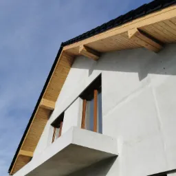 Surowa elewacja domu w trakcie budowy z drewnianym podbitką dachu i oknami z brązowymi ramami, widok z dołu na tle błękitnego nieba.