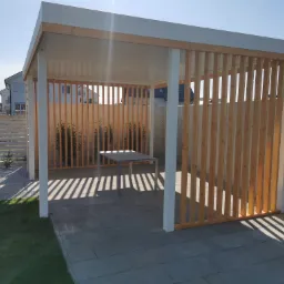 Nowoczesna, minimalistyczna konstrukcja z białych słupów i drewnianych listew, z płaskim dachem, na szarym brukowanym patio, z widokiem na domy w tle.