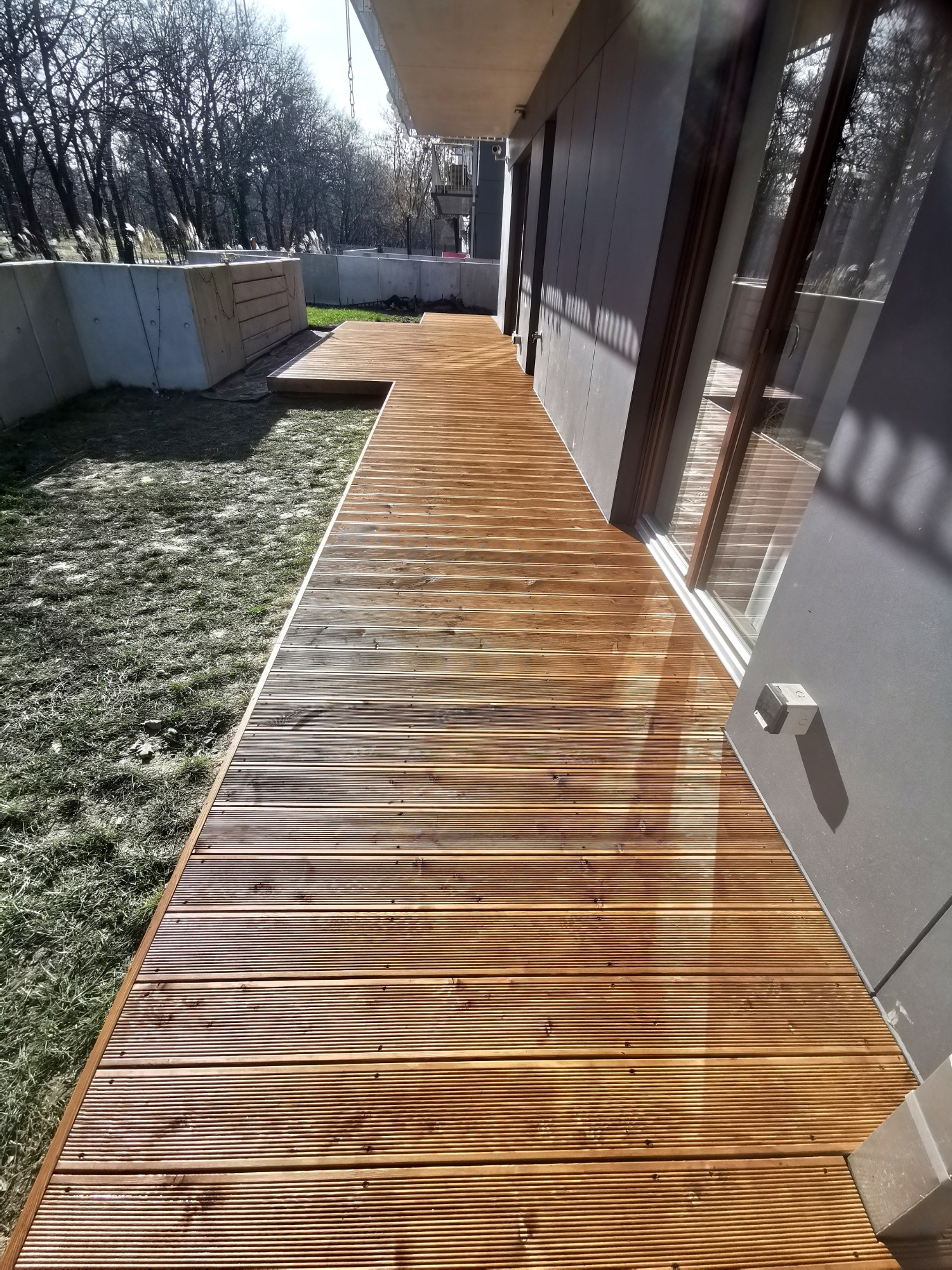 Nowoczesny taras z desek ułożonych wzdłuż budynku, z widoczną strukturą drewna i subtelnym oświetleniem słonecznym padającym na powierzchnię.