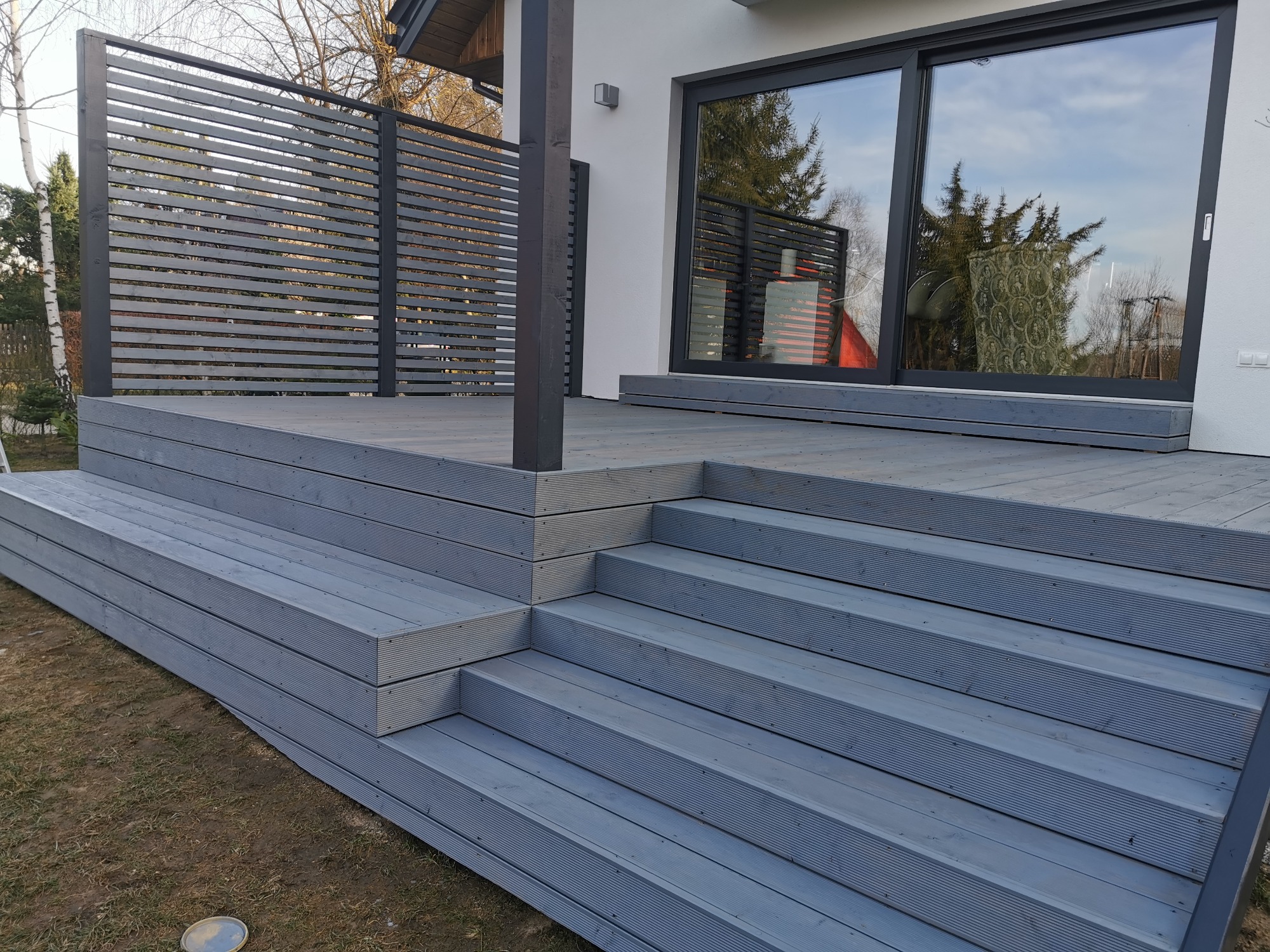 Szara, nowoczesna konstrukcja tarasu z widocznymi schodami i ażurową ścianką, przylegająca do białego budynku z dużymi przesuwnymi oknami.